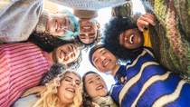 A diverse group of people stand close together in a circle, looking down at the camera. A diverse group of people stand close together in a circle, looking down at the camera.