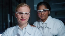 Two female scientists wearing white lab coats with the BASF logo and safety goggles stand in the laboratory and look into the camera.