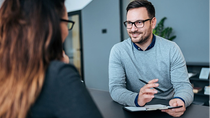 Zwei Personen sitzen an einem Tisch in einem modernen Büro und führen ein Gespräch, während sich die eine Person Notizen macht.
