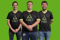 Drei Personen stehen nebeneinander vor einem grünen Hintergrund und tragen schwarze T‑Shirts mit BASF‑Logo im Gaming‑Design; die Personen präsentieren sich als Teil eines E‑Sports Teams.