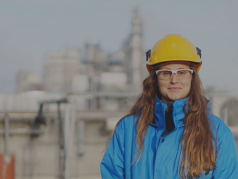 An engineer wearing a hard hat and safety goggles stands in front of an industrial plant. She is working on sustainable chemical innovations.