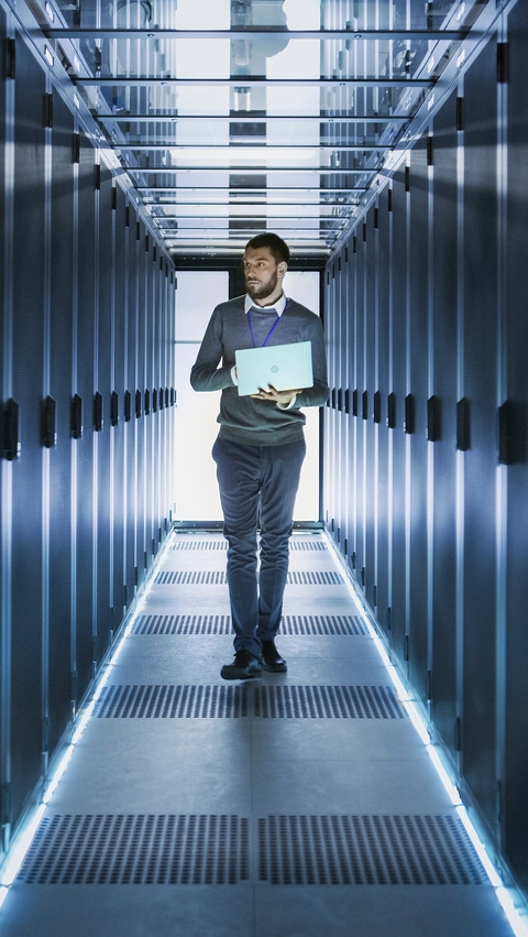 A technician carrying a laptop walks through a modern data center surrounded by illuminated server racks.
